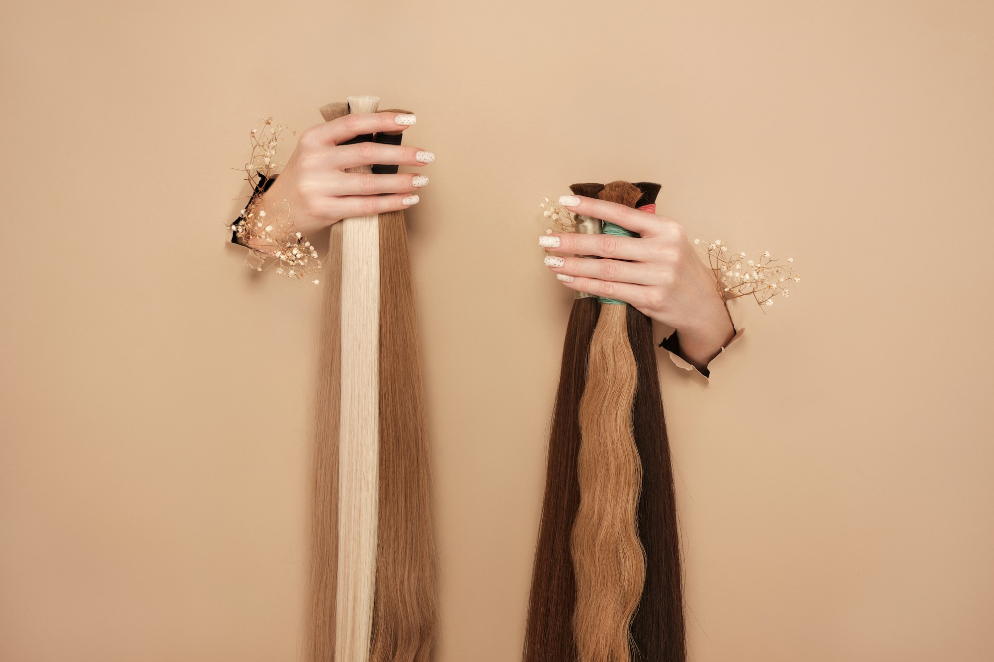 Hands with flowers hold strands of hair for extensions on a beige background.